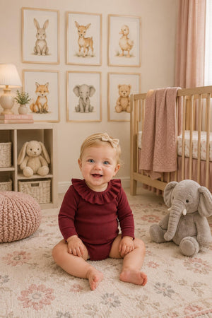 A happy baby girl sits on a soft rug, wearing a burgundy ribbed long-sleeve bodysuit with ruffles. The cozy nursery features animal artwork and plush toys, creating a warm atmosphere.