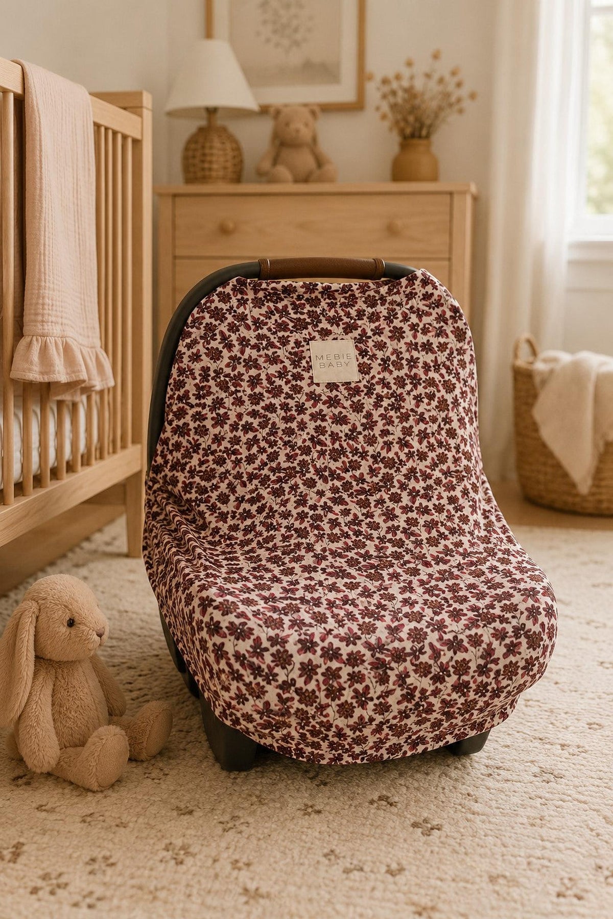 A floral-patterned multi-use cover in shades of burgundy and cream drapes over a car seat, set in a cozy nursery with a wooden crib and soft toys nearby.