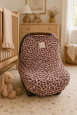 A floral-patterned multi-use cover in shades of burgundy and cream drapes over a car seat, set in a cozy nursery with a wooden crib and soft toys nearby.