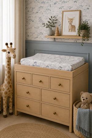 A wooden changing table with multiple drawers features a muslin changing pad cover in a soft, neutral color. A giraffe and teddy bear plush toys complement the cozy nursery setting.