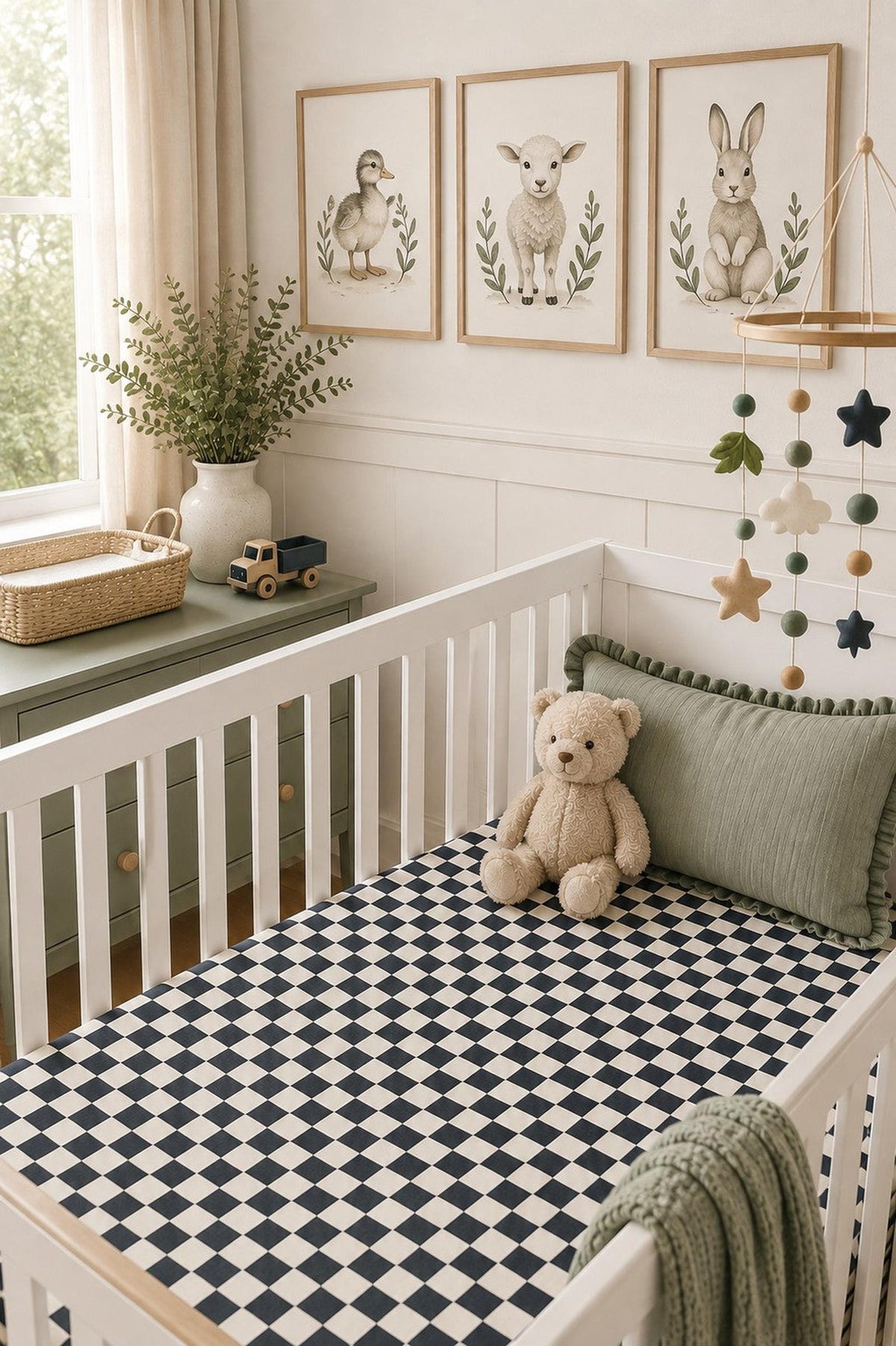 A cozy nursery features a white crib with a charcoal checkered muslin crib sheet, soft teddy bear, and decorative pillows. Framed animal prints and green accents create a serene atmosphere.