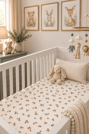 A cozy nursery features a crib with a cream floral muslin sheet, surrounded by soft toys and framed animal art. The gentle colors create a warm, inviting atmosphere for babies.