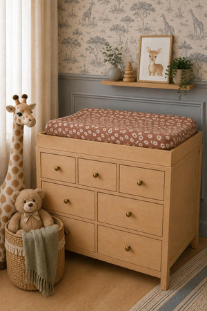 A wooden changing table with eight drawers features a brown floral changing pad cover. Nearby, a plush teddy bear sits in a woven basket next to a giraffe decoration.