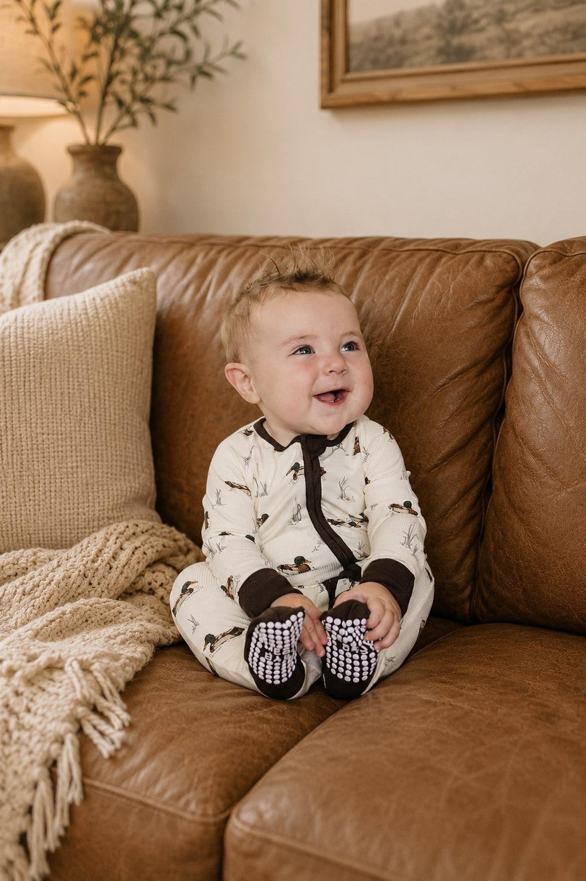 A baby sits on a brown leather couch, wearing a cream-colored ribbed bamboo pajama with a playful duck print. The cozy setting features soft textures and warm lighting.