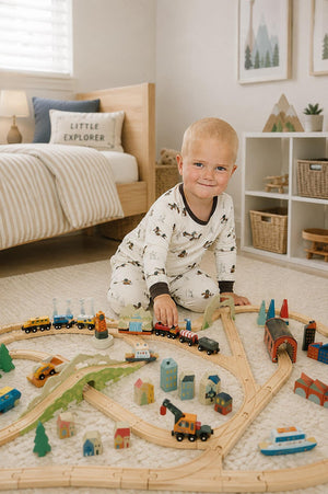 A young child in a cozy, ribbed bamboo pajama set plays on a wooden train track surrounded by colorful toy buildings and trees in a bright, softly decorated room.