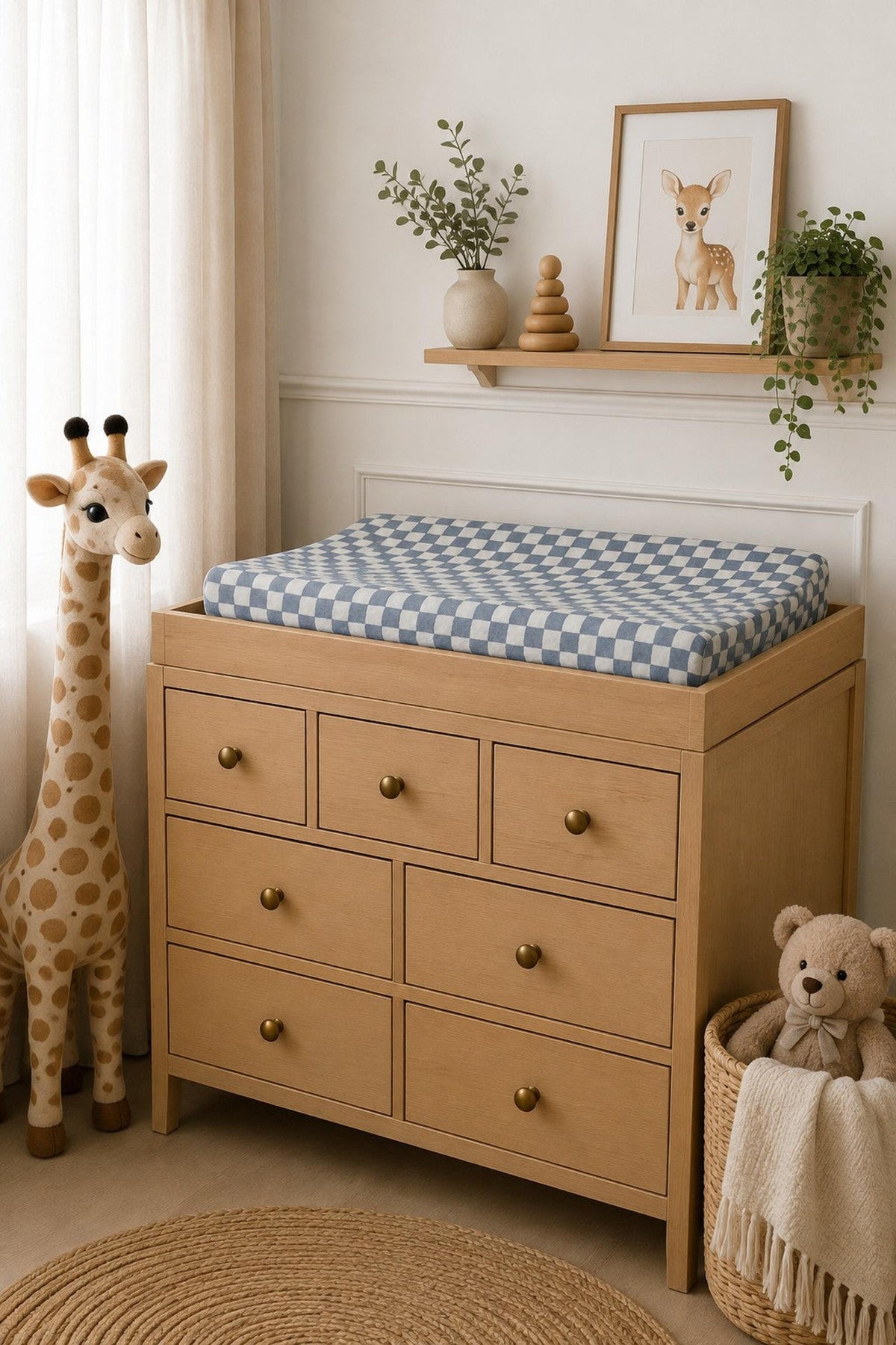 A changing table features a dusty blue wavy checkered muslin changing pad cover on top, alongside a plush giraffe and teddy bear, creating a cozy nursery atmosphere.