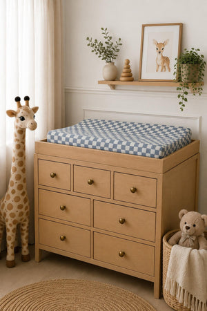A changing table features a dusty blue wavy checkered muslin changing pad cover on top, alongside a plush giraffe and teddy bear, creating a cozy nursery atmosphere.
