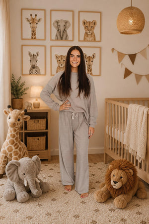 A woman stands in a cozy nursery, wearing a soft gray loungewear set made from breathable cotton. The room features animal-themed decor, a crib, and plush toy elephants and lions.