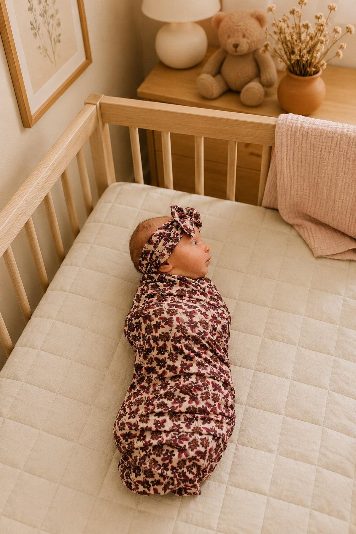 A newborn is peacefully swaddled in an antique floral ribbed bamboo stretch swaddle, lying on a soft, quilted crib mattress.