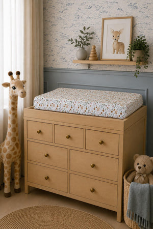 A wooden changing table features a floral muslin changing pad cover, surrounded by a soft, neutral nursery decor including a giraffe plush toy and framed artwork.
