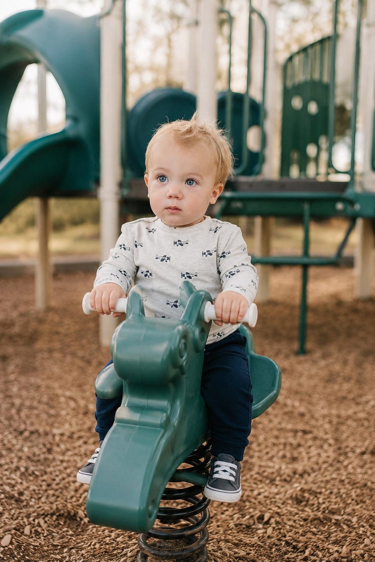A toddler sits on a green spring rocker at a playground, wearing a light gray shirt with a car pattern and navy pants.