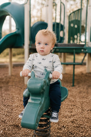 A toddler sits on a green spring rocker at a playground, wearing a light gray shirt with a car pattern and navy pants.