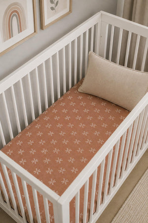 A cozy crib features a soft, peach-colored muslin sheet adorned with a delicate floral pattern. The minimalist white crib is complemented by a neutral-toned pillow and styled artwork in the.