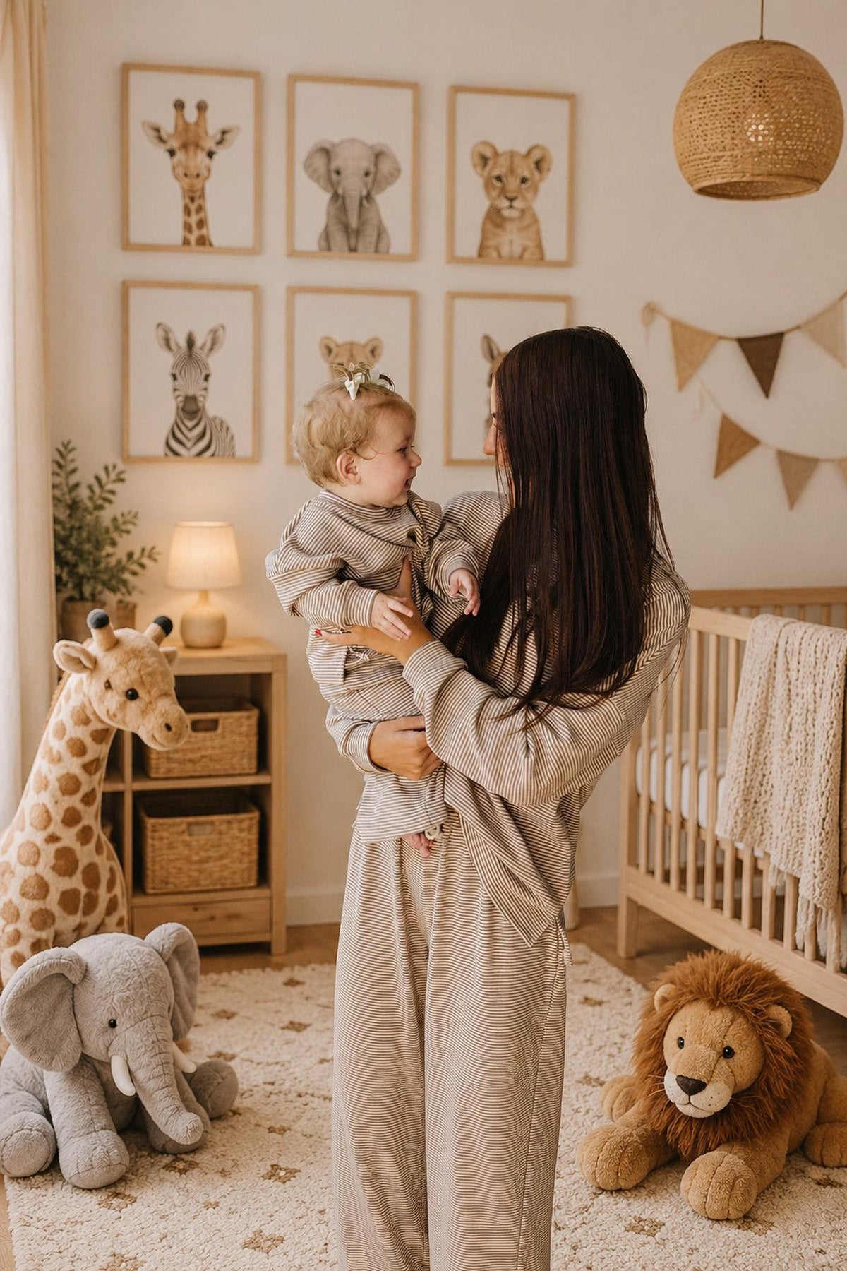A mother holds her smiling toddler in a cozy nursery adorned with animal-themed artwork and plush toys. They both wear matching striped cotton loungewear, creating a warm family atmosphere.