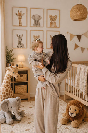A mother holds her smiling toddler in a cozy nursery adorned with animal-themed artwork and plush toys. They both wear matching striped cotton loungewear, creating a warm family atmosphere.
