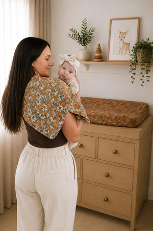 A woman holds a baby in a cozy nursery, featuring a floral muslin changing pad cover in warm tones.
