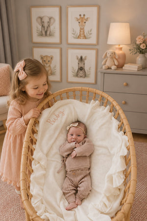 A young girl smiles while standing beside a cozy rattan bassinet. Inside, a newborn dressed in a soft mauve knit outfit rests on a plush white blanket.