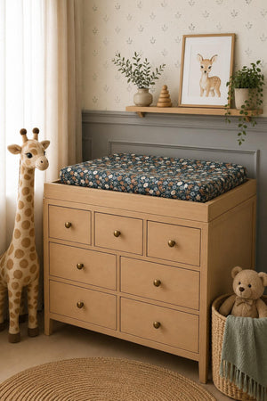 A changing pad covered with a midnight floral print muslin cover sits atop a light wood dresser.