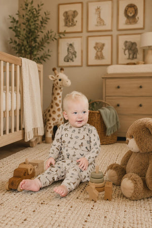 A smiling baby sits on a soft rug surrounded by wooden toys, wearing a light gray outfit with cactus prints.