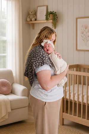 A woman in a cozy, light-filled nursery holds a swaddled baby against her chest, wearing a floral-patterned shawl. The room features soft decor, including a crib and plush furnishings.