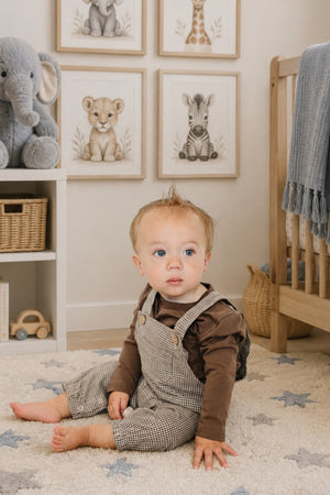 A toddler sits on a soft, star-patterned rug in a nursery, wearing brown plaid cotton overalls and a brown long-sleeve shirt, surrounded by animal-themed artwork and plush toys.