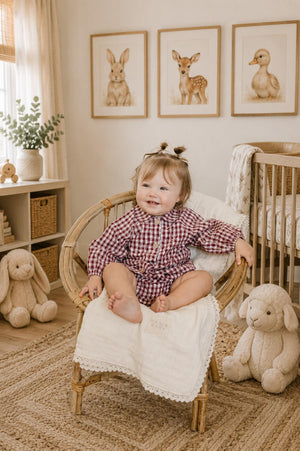 A cheerful baby girl sits in a rattan chair, wearing a red gingham bloomer set with a Peter Pan collar. Soft toys and framed animal prints decorate the cozy room.