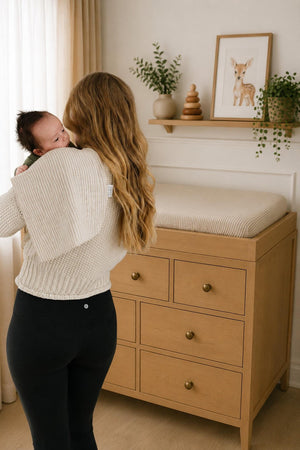 A mother cradles her baby while standing near a wooden changing table, adorned with a light taupe striped muslin changing pad cover. The room features neutral tones.