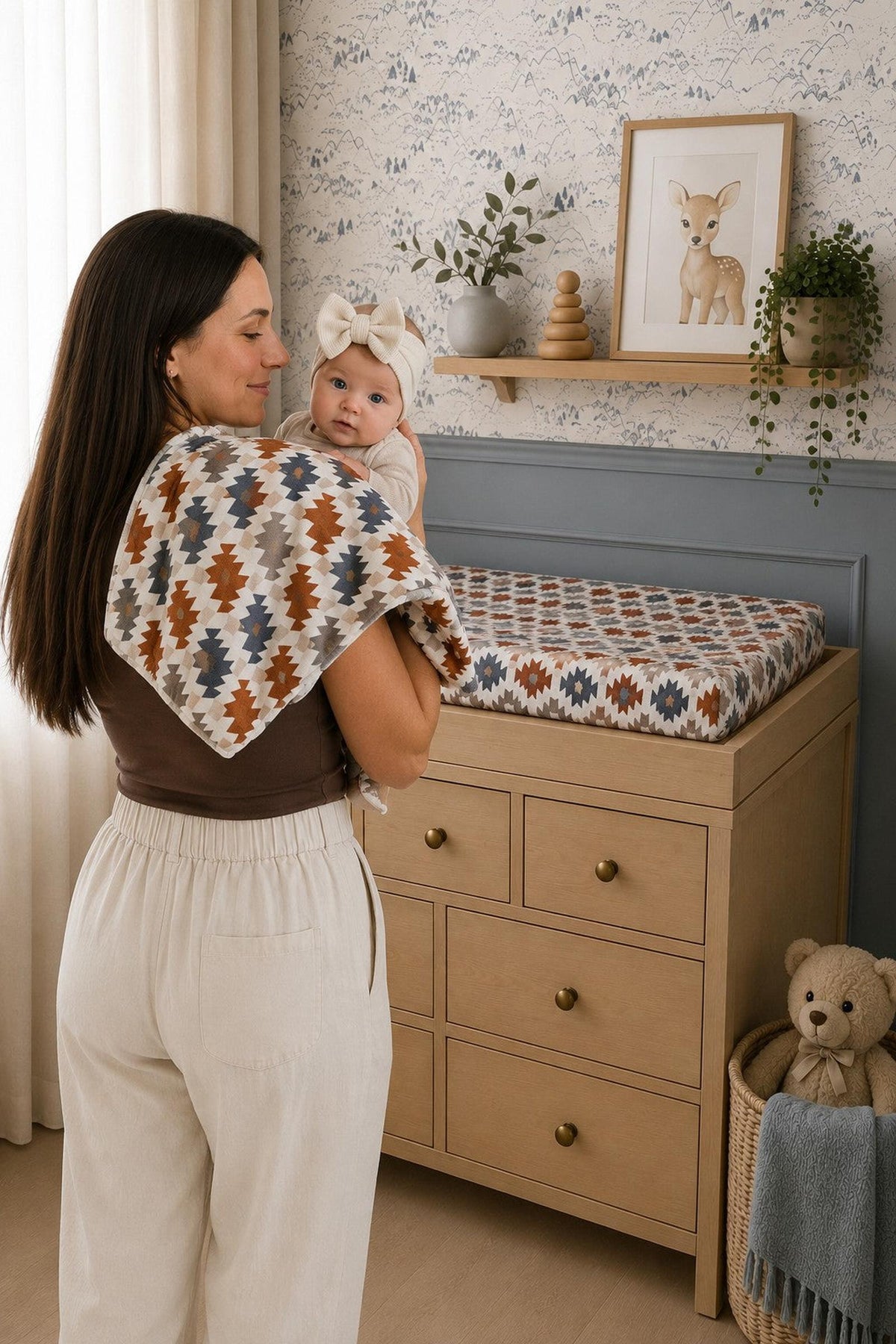 A woman holds a baby while standing near a changing table topped with a Western Aztec muslin changing pad cover in earthy tones.