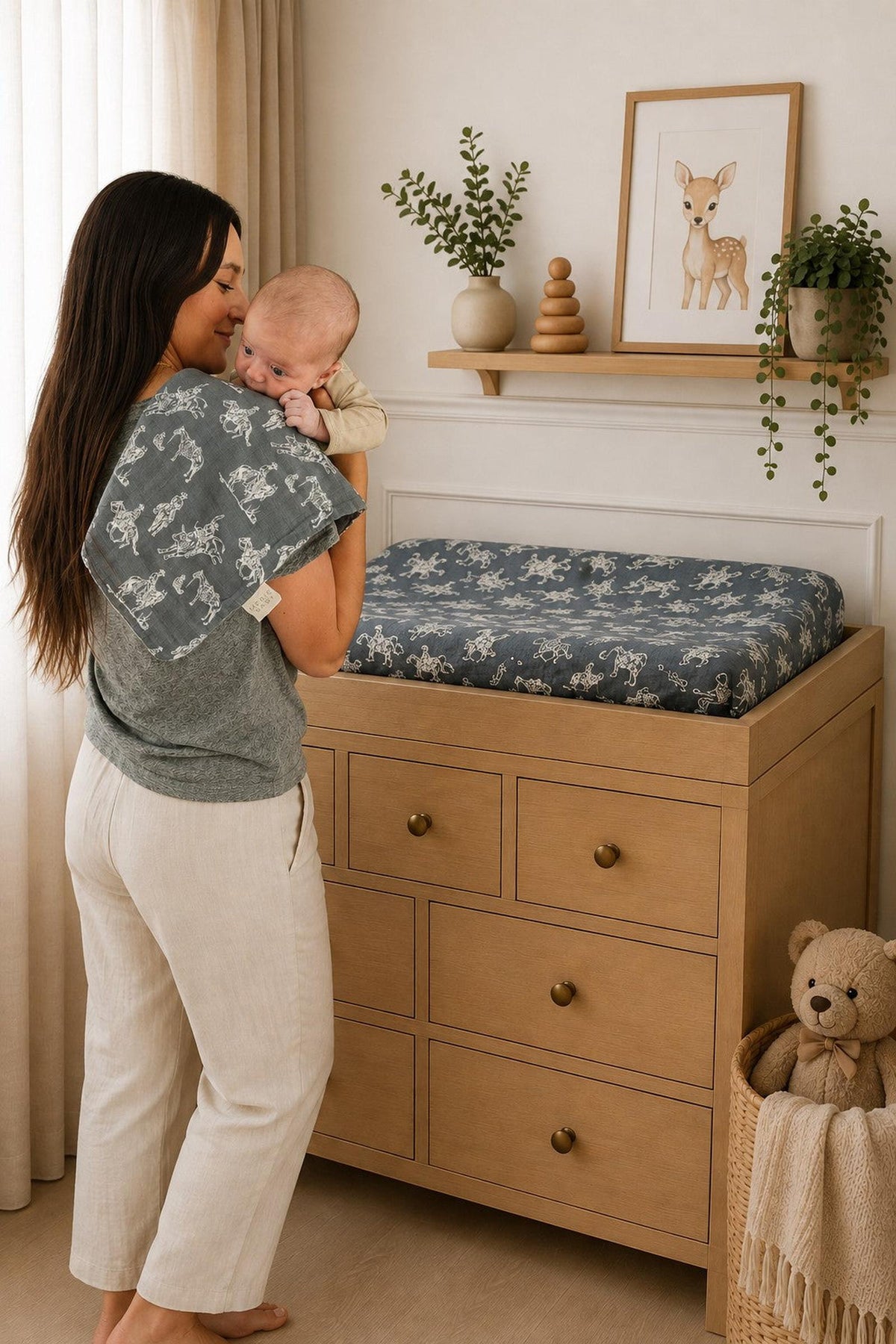 A mother holds her baby while standing next to a changing table topped with a Wild West muslin changing pad cover featuring a rodeo print.