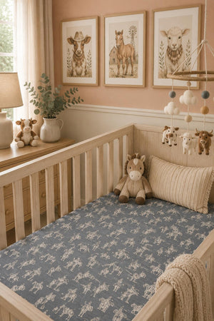 A cozy nursery features a wooden crib with a blue Wild West muslin crib sheet, adorned with rodeo designs. Soft toys and framed animal prints add warmth to the space.