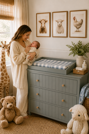 A mother in a cozy setting holds a baby near a blue changing pad cover on a dresser, surrounded by soft toys and framed animal pictures, conveying a gentle nursery.