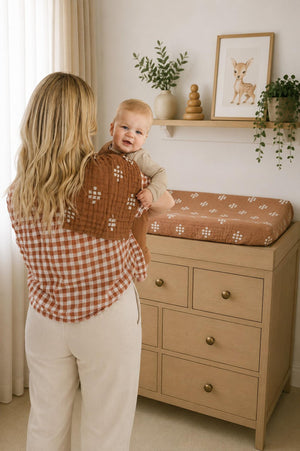 A parent holds a smiling baby while standing near a changing table adorned with a rust-colored muslin changing pad cover. The room features soft lighting, greenery,.