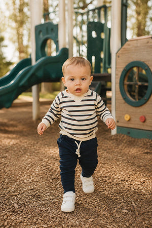 A toddler in a striped crewneck sweatshirt and navy pants walks on a playground covered in wood chips, with green slides and play structures in the background, enjoying outdoor fun.