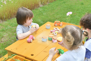 Three children play at a wooden picnic table outdoors, engaging with colorful toy vehicles and food items. The grassy setting features blooming flowers, creating a vibrant, playful atmosphere.