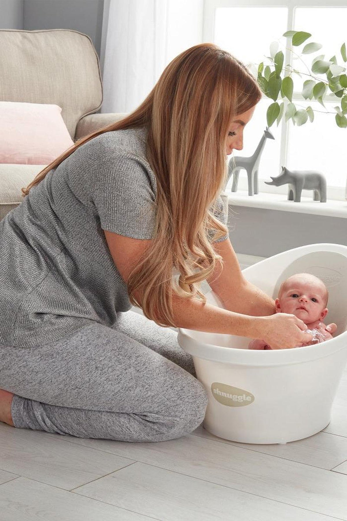 A woman in grey clothing bathes a baby in a white Shnuggle Baby Bath in a cozy room.