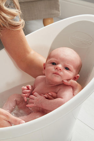 A newborn is bathed in a white Shnuggle baby bath, supported by a caregivers hand in a cozy setting.