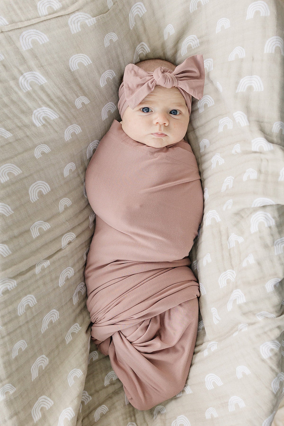 A baby is wrapped in a dusty rose bamboo swaddle with a matching headband, lying on a patterned blanket.