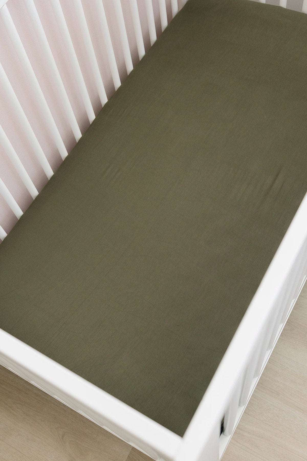 Olive-colored crib sheet on a white crib with wooden flooring, showcasing a simple and elegant nursery setting.