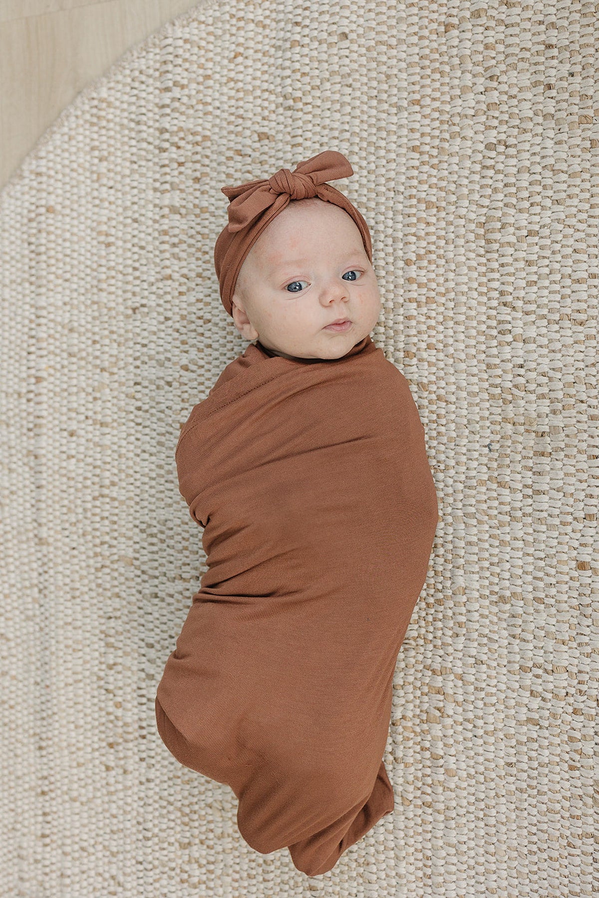 A baby is wrapped in a rust-colored swaddle blanket with a matching head wrap, lying on a textured rug.