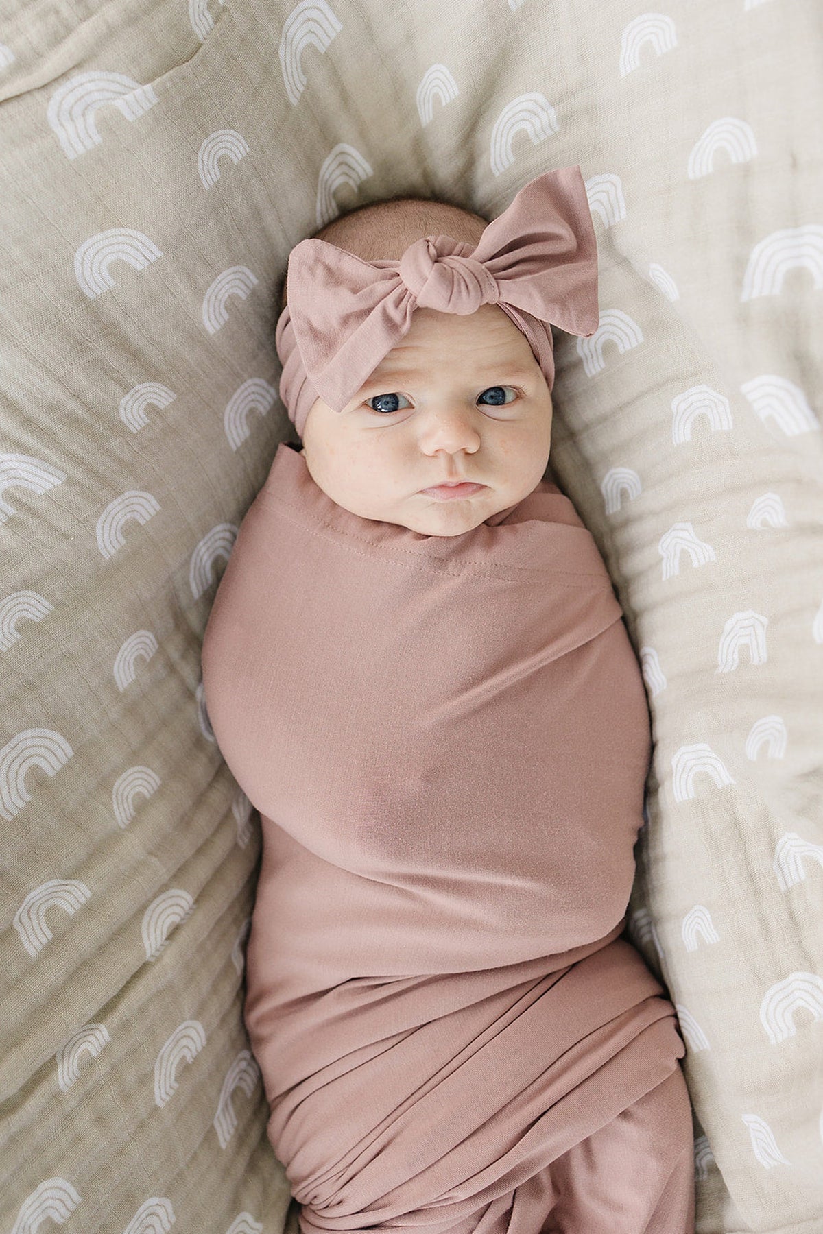 A baby wrapped in a dusty rose bamboo swaddle with a matching head wrap, lying on a rainbow-patterned blanket.