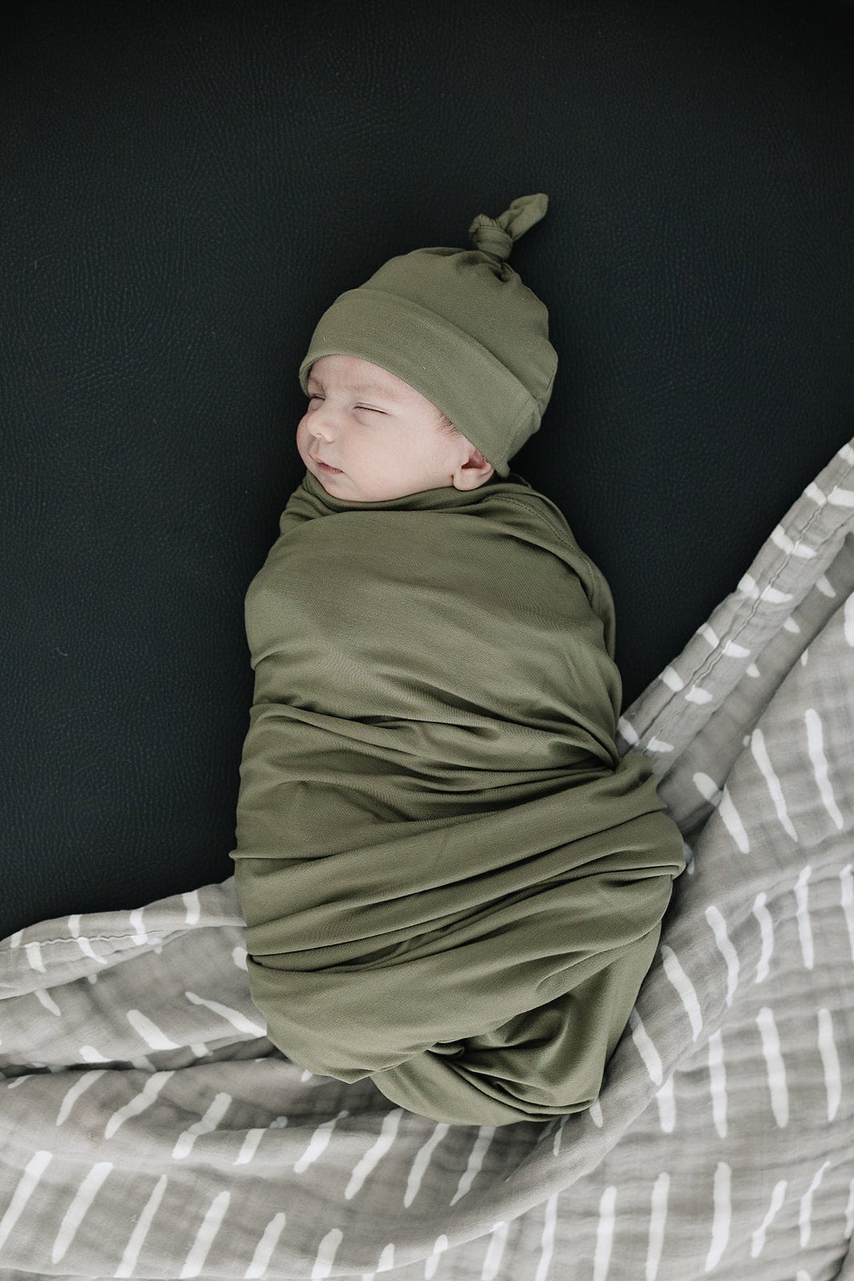 A newborn sleeps peacefully in an olive green swaddle and knot hat, lying on a patterned blanket.