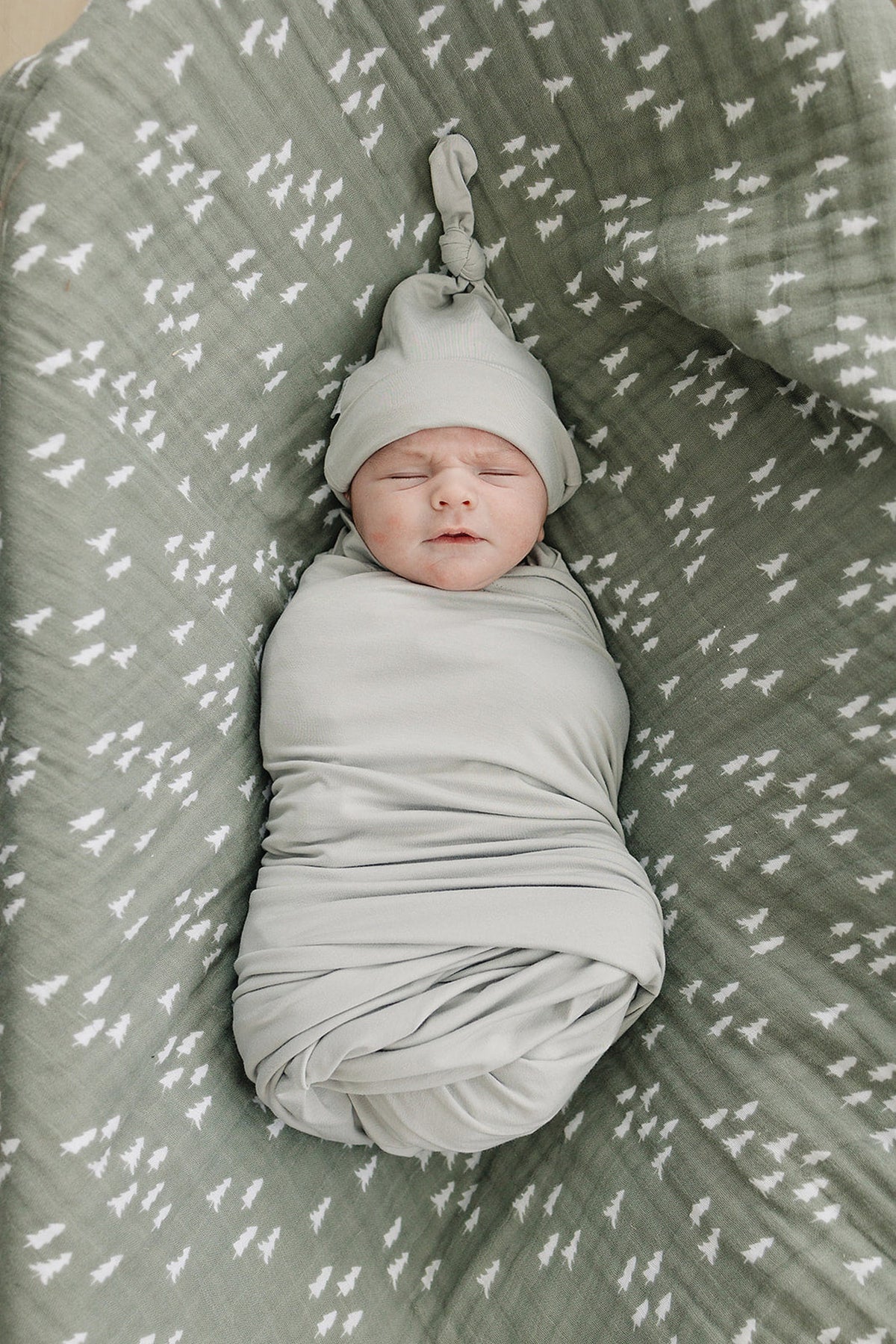 Newborn in a sage green knot hat and swaddle, lying on a patterned blanket with tree designs.