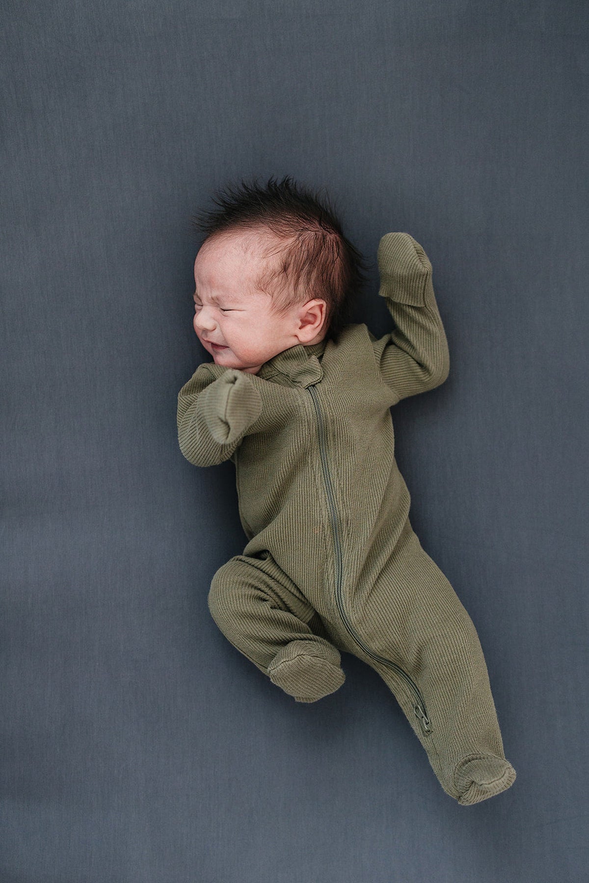 A baby wearing a green ribbed zipper footed pajama, lying on a dark gray background.