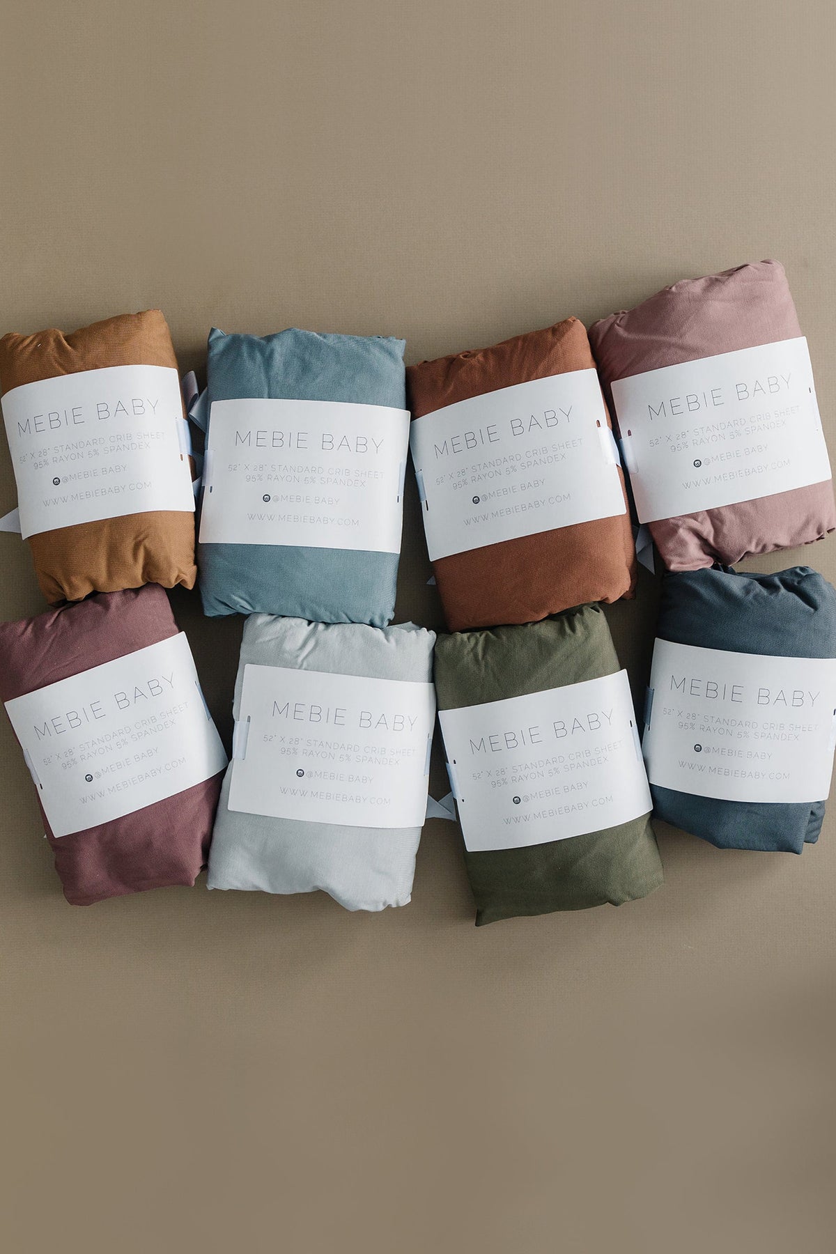 Seven Mebie Baby crib sheets in various colors, neatly folded with labels, displayed on a neutral background.