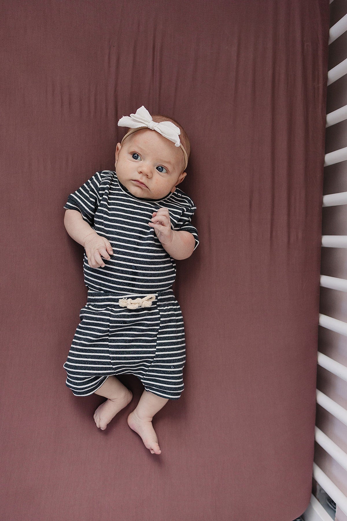 A baby in striped outfit and headband lies on a plum-colored crib sheet.