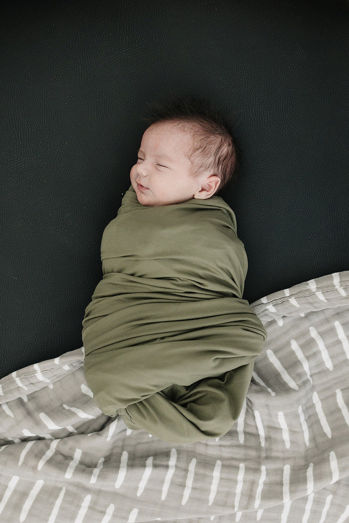 A baby is swaddled in an olive green blanket, lying on a dark surface with a patterned gray blanket.