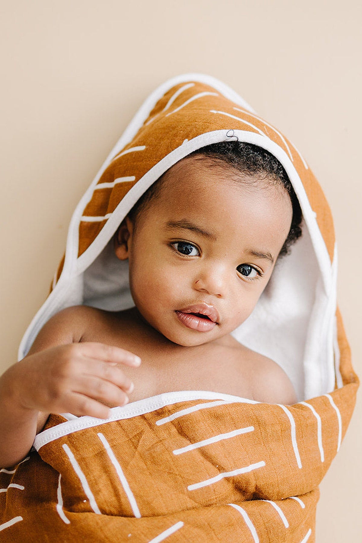 A baby wrapped in a mustard-colored hooded towel with white stripes, lying on a soft beige surface.
