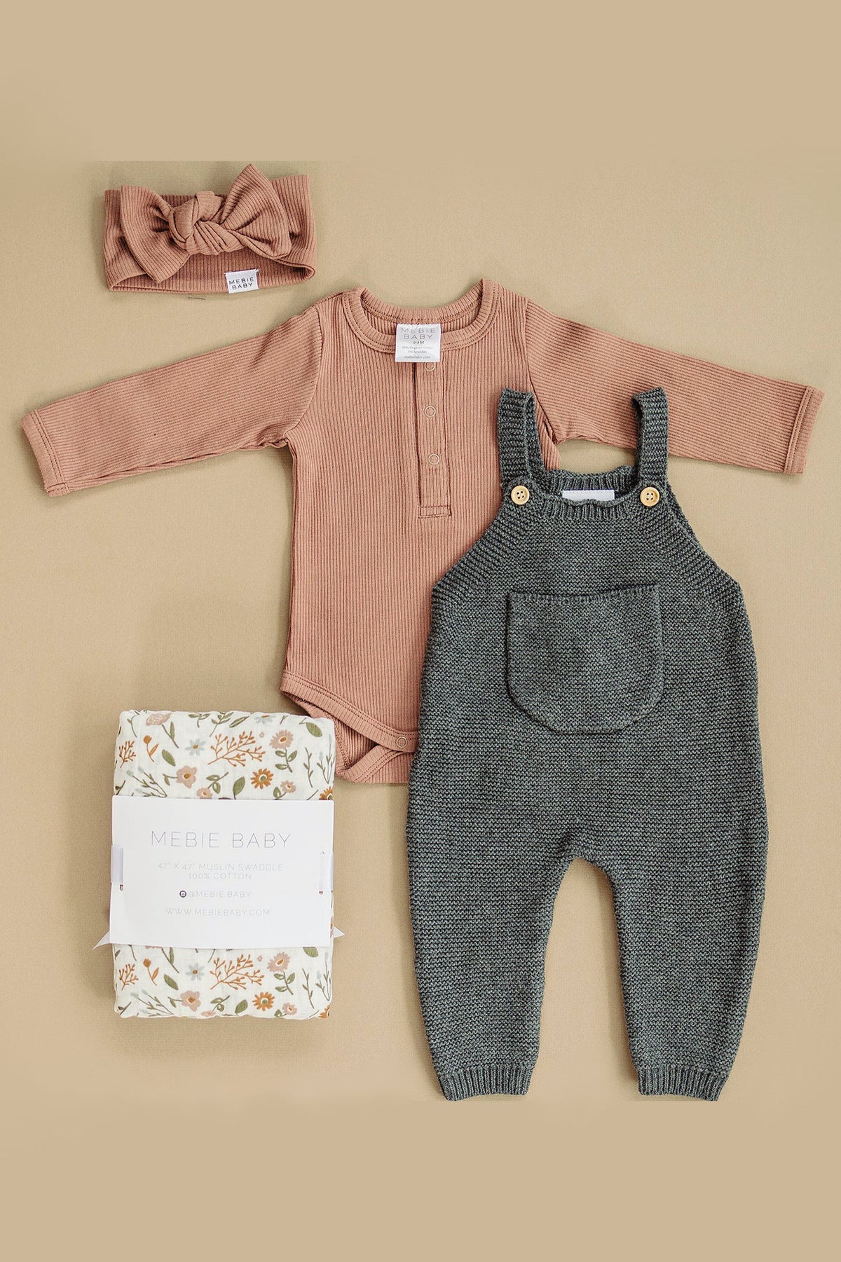 Dusty rose long-sleeve bodysuit, gray knit overalls, floral swaddle, and matching headband on a beige background.
