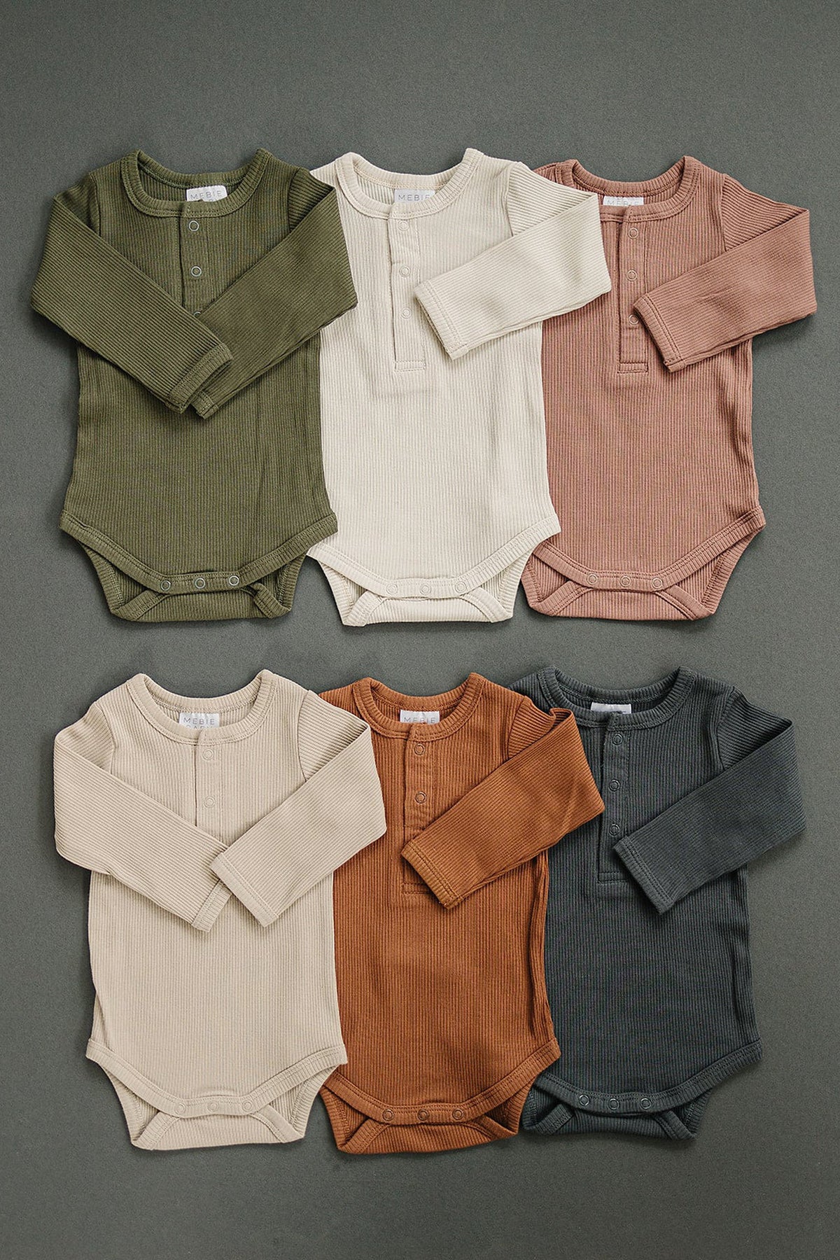 Six ribbed long sleeve baby bodysuits in earthy tones, featuring snaps, made from organic cotton.