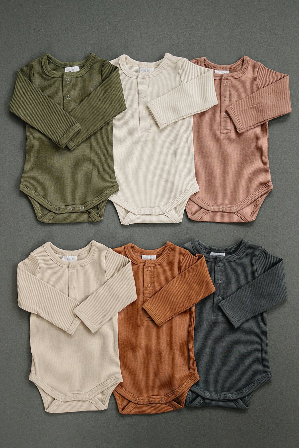 Six ribbed long sleeve baby bodysuits in olive, cream, pink, beige, rust, and charcoal, displayed on a gray background.